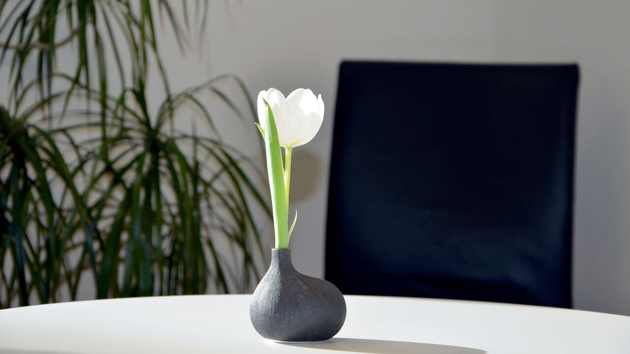 Eine einzelne Blume mit weißen Blütenblättern steht in einer grauen Vase auf einem Tisch.